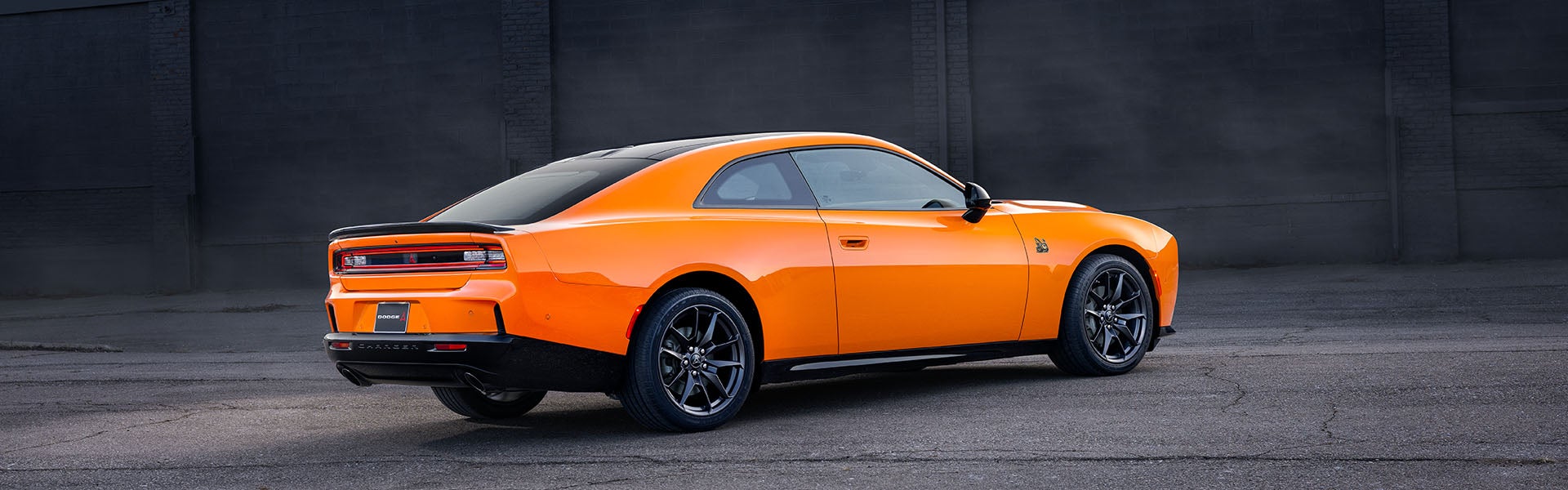 Rear three-quarter view of a bright orange electric Dodge Charger parked on asphalt, against a dark brick wall.