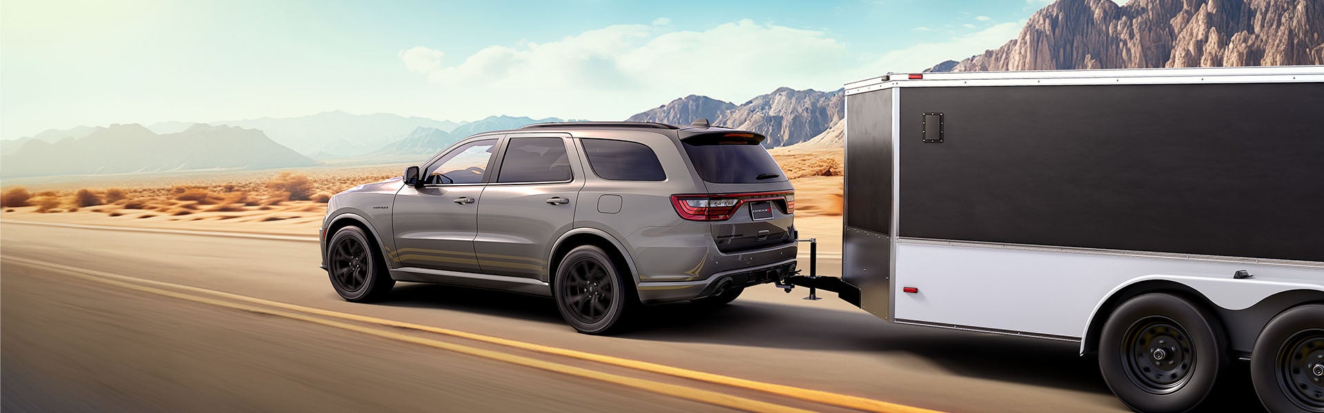 Gray SUV towing a large black and white enclosed trailer on a highway through a desert landscape with mountains.