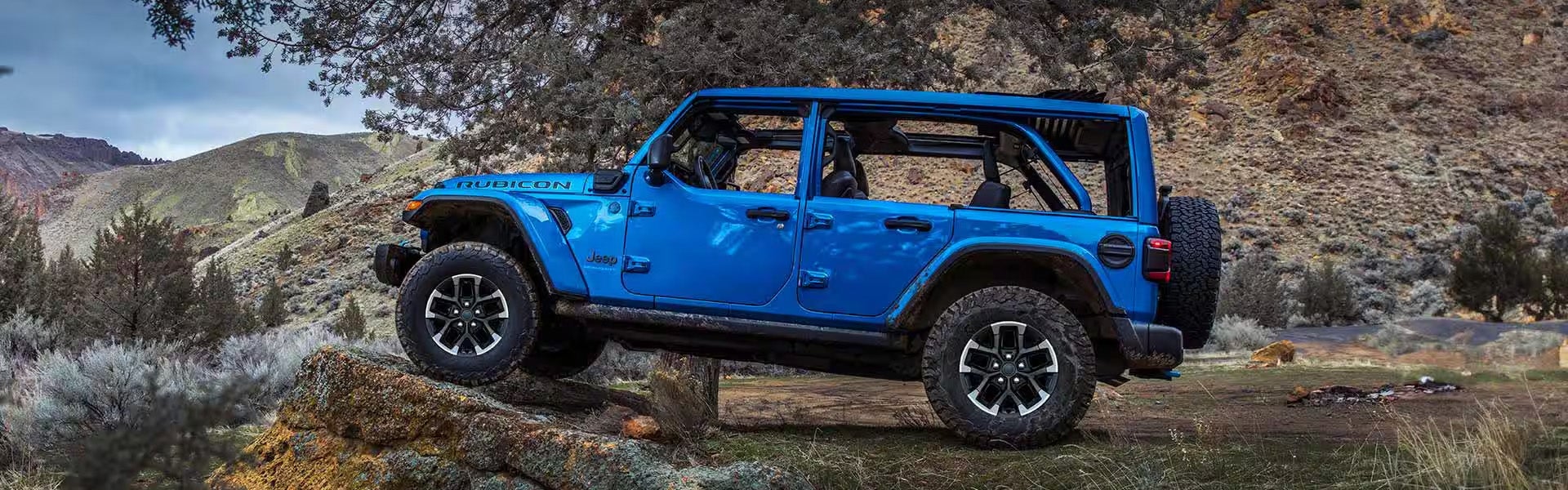 A blue Jeep Wrangler Rubicon with its top off, angled on a rocky hill with an open desert landscape.