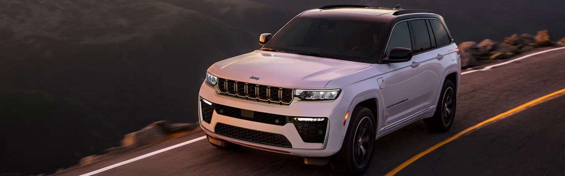 White Jeep Grand Cherokee 4xe SUV driving on a winding road with dark mountains in the background.