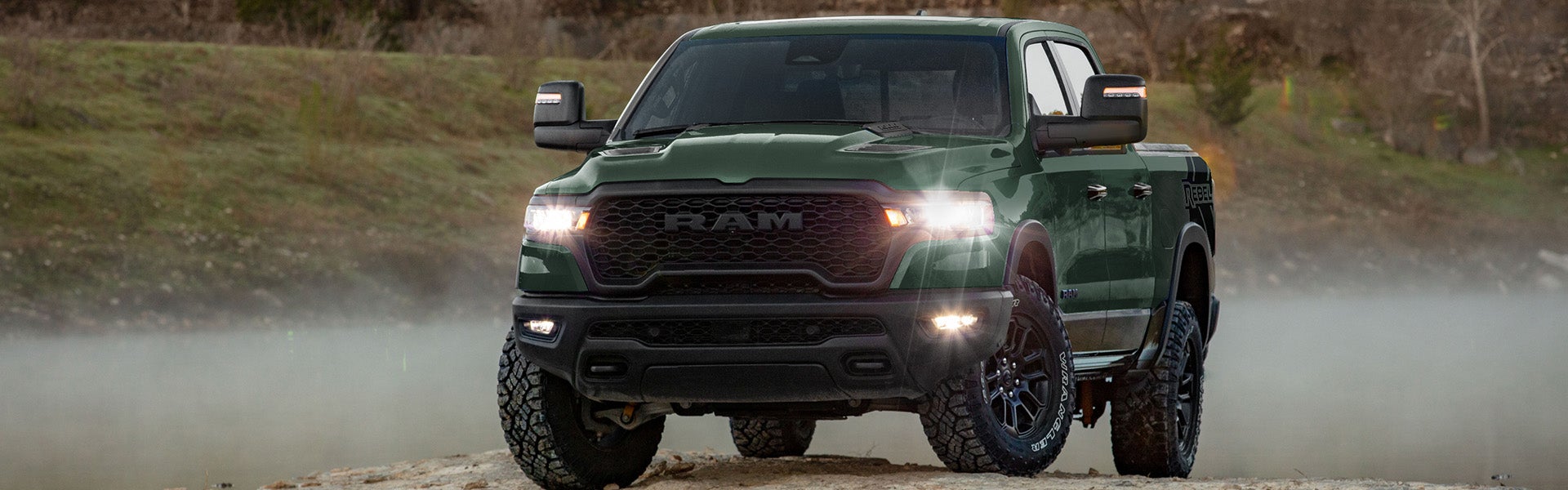 A dark green Ram pickup truck with headlights on, parked on a muddy, rocky terrain surrounded by brush and hazy air.