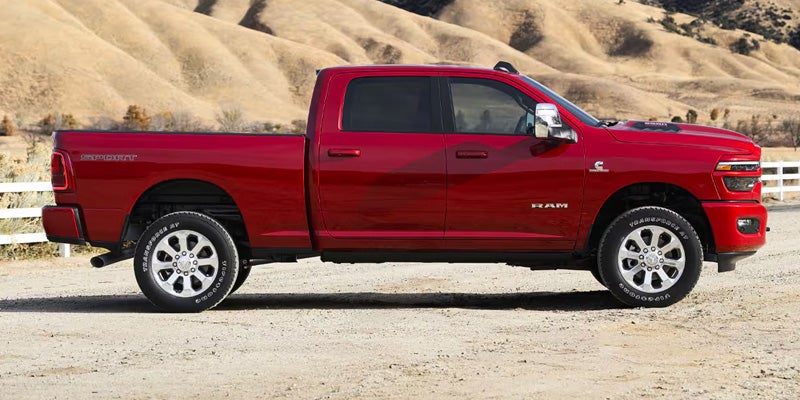 Side view of a red Ram pickup truck with a four-door crew cab, parked on a dirt road with hills in the background.