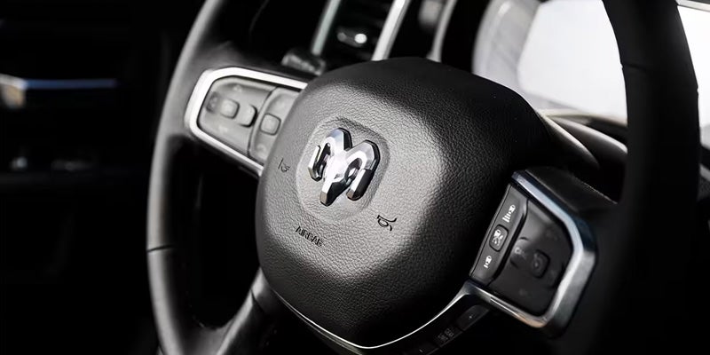 A close-up of a black car steering wheel, featuring the shiny chrome Ram logo and silver-trimmed control buttons.