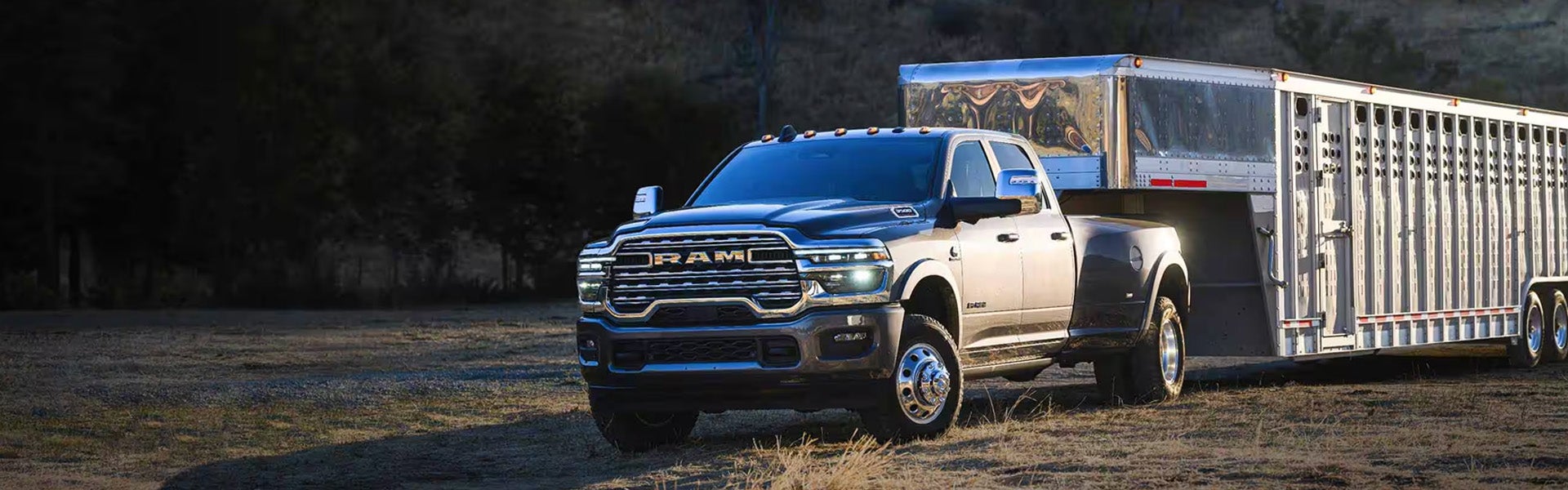 A grey 2026 RAM 3500 pickup truck pulling a large, silver livestock trailer across a grassy field at dusk.