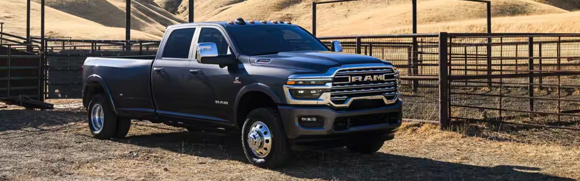 A dark gray Ram 3500 heavy-duty pickup truck with chrome accents, parked on dirt in front of farm corrals.