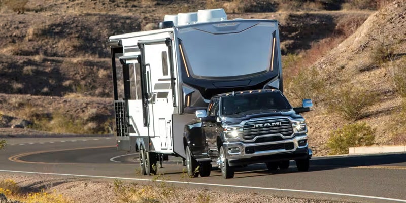 A dark gray Ram 3500 truck with a silver grille pulls a large white and dark gray RV on a curved desert road.
