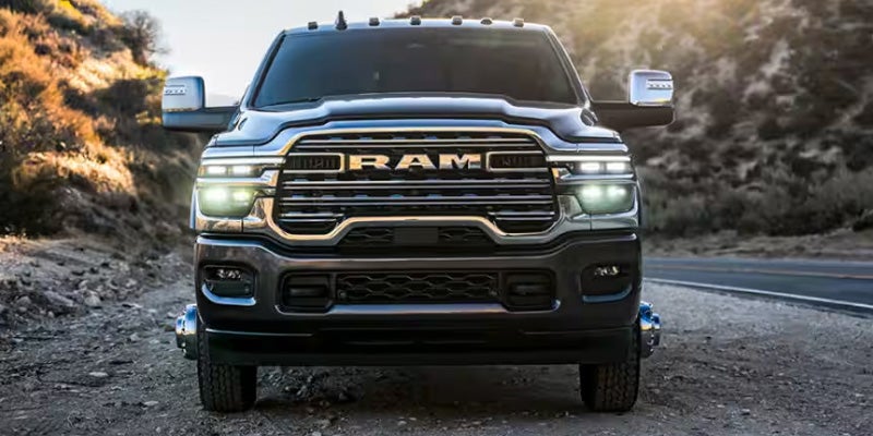 Front view of a dark gray Ram 3500 pickup truck with headlights on, parked on a dirt road next to pavement and hills.