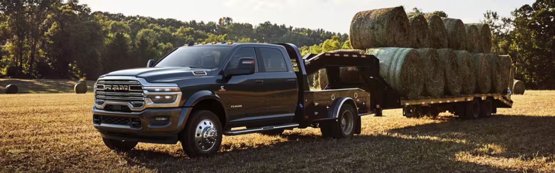 A dark gray Ram truck with a flatbed trailer full of hay bales on a field, trees in the background.